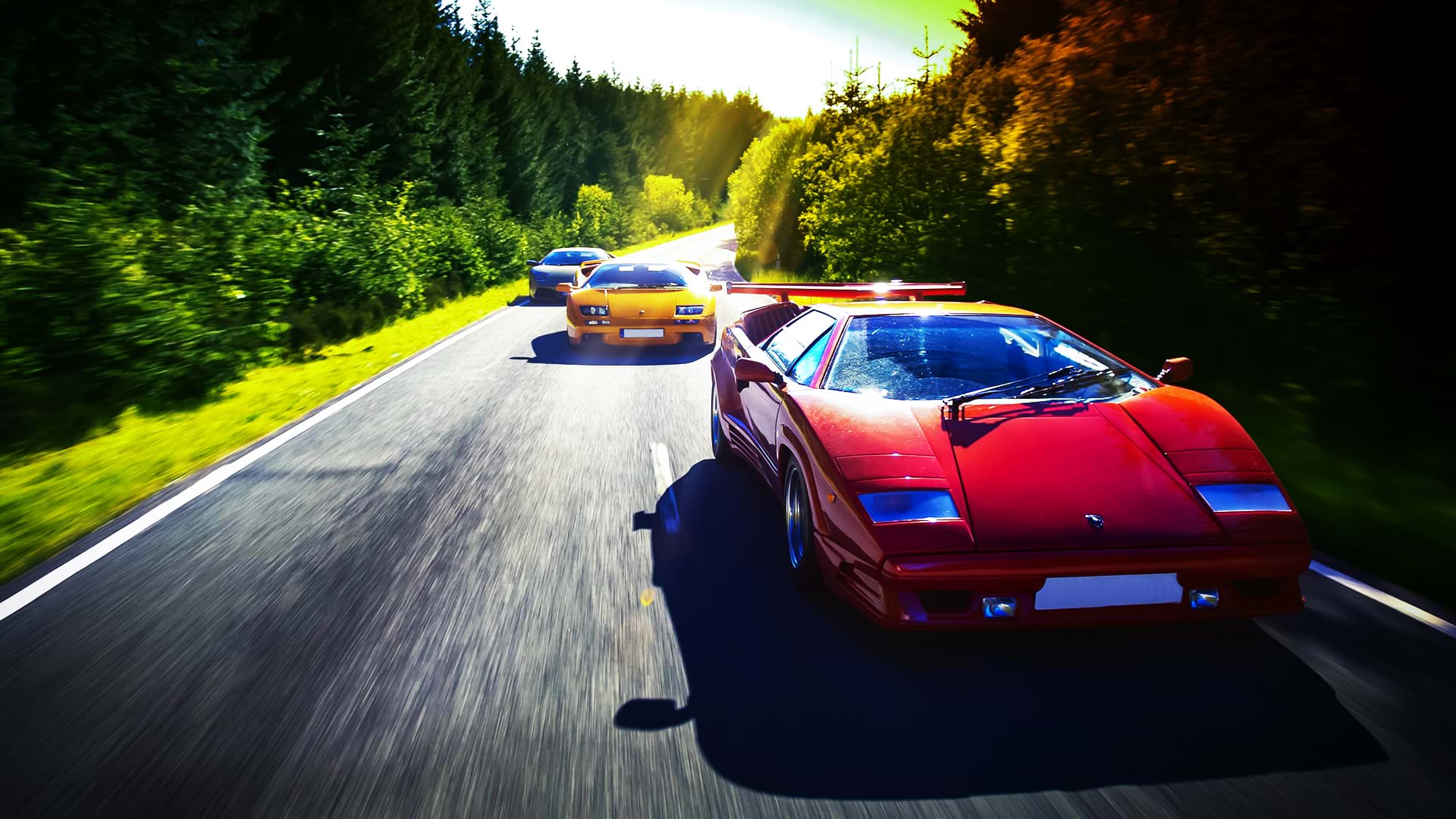 A colorful Lamborghini convoy: Countach 25th Anniversary, Diablo 6.0, and Murciélago, races along a scenic road on a bright day.