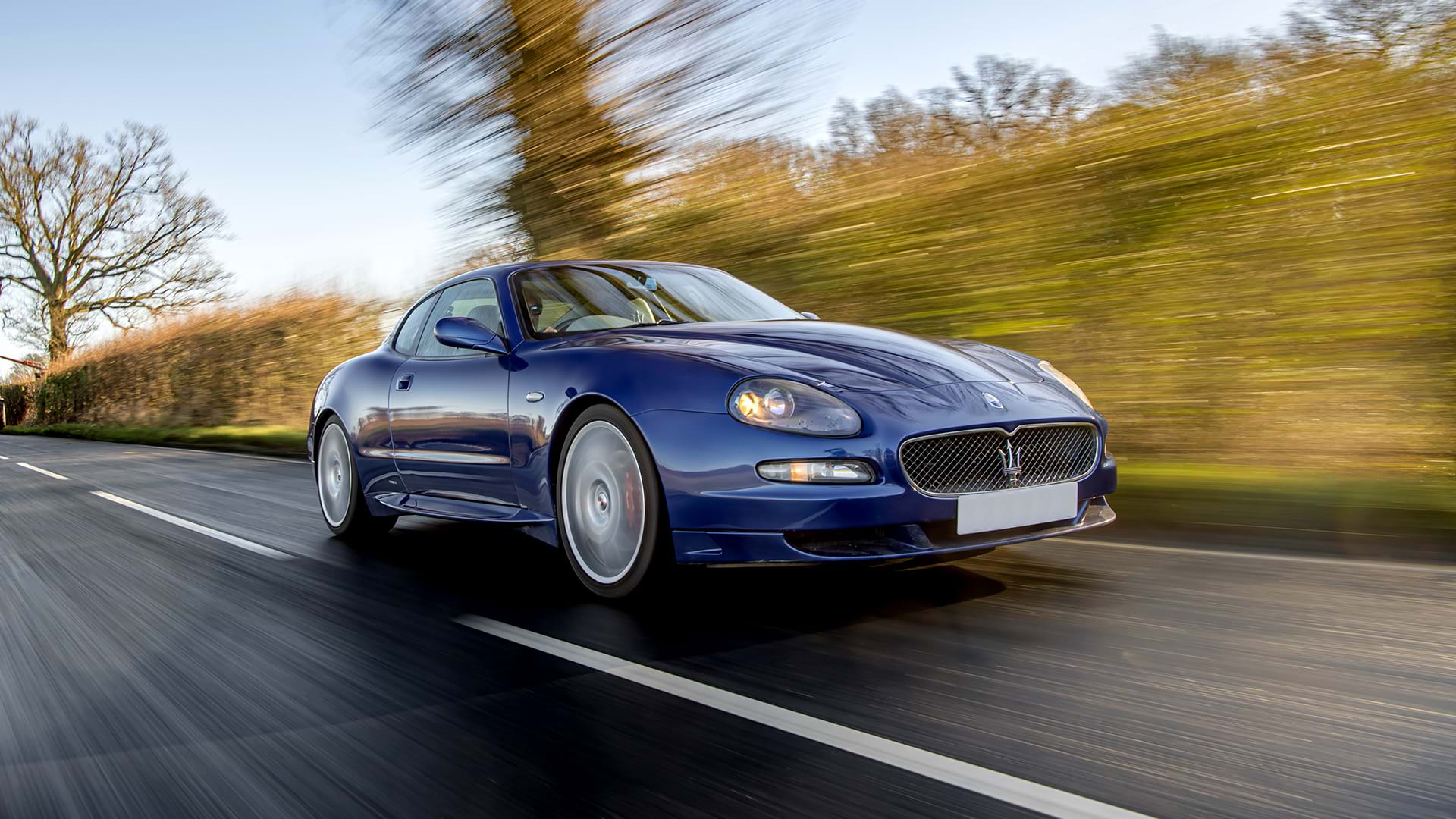 Classic Blue Maserati GranSport driving along a country road with blue skies overhead.