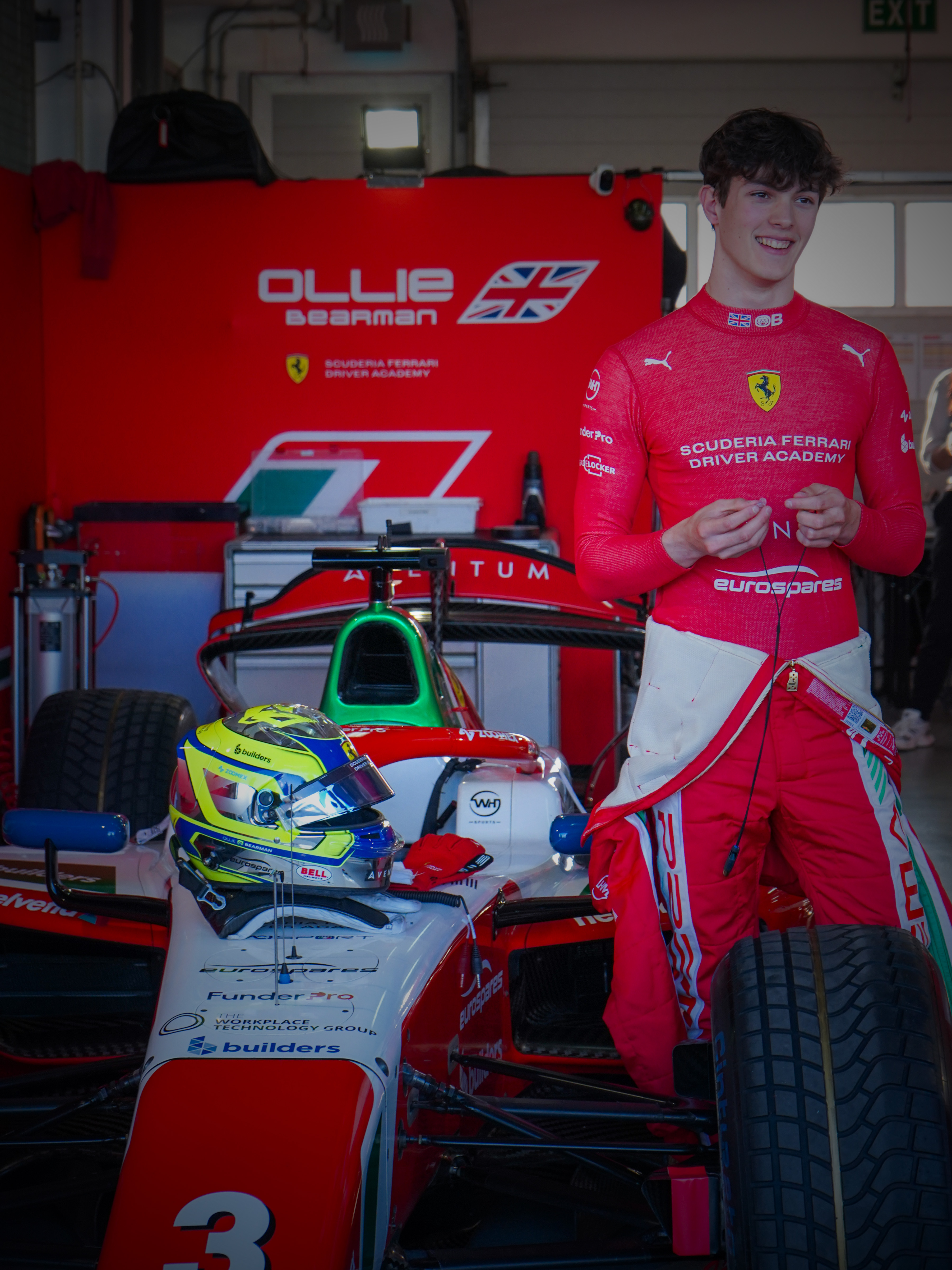 Image of young racing driver Oliver Bearman standing next to his Formula 2 car in the Prema Racing team garage at the 2024 Bahrain F2 race weekend. He is wearing his racing suit with the Eurospares sponsor logo visible on his shirt.